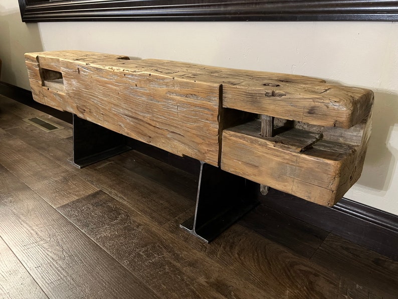 May include: A rustic wooden bench with a weathered, natural finish. The bench is supported by two black metal legs. The wood has visible knots and imperfections, adding to its unique character. The bench is a substantial piece of furniture.