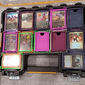 May include: A black and yellow plastic storage case with multiple compartments, each holding trading cards. The cards feature colorful illustrations and text. Some compartments are empty, while others hold cards in purple, green, and orange frames.