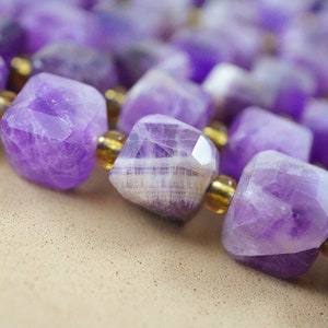 May include: A close-up of a purple amethyst gemstone necklace. The necklace is made of faceted, square-shaped beads with a gold-toned spacer bead between each amethyst bead.