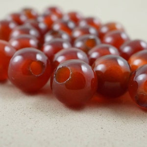 May include: A close-up of a pile of smooth, round, red carnelian beads. The beads have a hole drilled through the center for stringing.