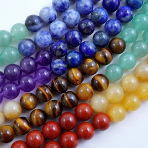 May include: A set of seven chakra gemstone beads in various colours, including green, blue, purple, brown, yellow, and red. The beads are round and smooth, and they are arranged in a row.