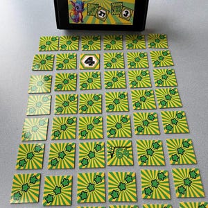 May include: A memory game with green and yellow square cards featuring a sunburst design and a three-leaf clover. The game box reads "cubes memory" and has a cartoon dragon. Some cards have numbers and math symbols.