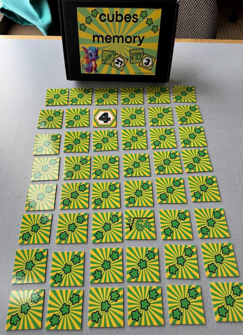 May include: A memory game with green and yellow square cards featuring a sunburst design and a three-leaf clover. The game box reads "cubes memory" and has a cartoon dragon. Some cards have numbers and math symbols.