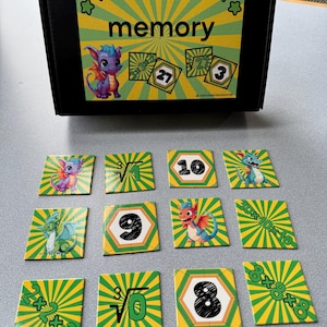 May include: A black box with the words "cubes memory" and a cartoon dragon. Twelve square cards with dragon illustrations and math problems are scattered in front of the box. The cards have green and yellow striped backgrounds.
