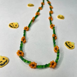 Könnte beinhalten: Eine grüne und orangefarbene Perlenkette mit kleinen blütenförmigen Perlen. Die Kette liegt auf einem weißen Hintergrund.