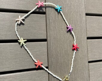 Peut inclure: Un collier de perles blanches avec des breloques en forme d'étoiles de mer colorées. Le collier a un fermoir doré.