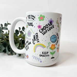 May include: White ceramic mug with a colorful design featuring flowers, rainbows, and positive affirmations like "Choose Self Love", "Trust Your Self", and "Keep Growing".