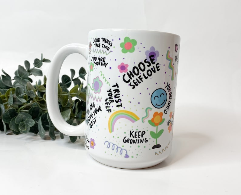 May include: White ceramic mug with a colorful design featuring flowers, rainbows, and positive affirmations like "Choose Self Love", "Trust Your Self", and "Keep Growing".