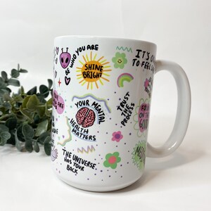 May include: White ceramic mug with a colorful design featuring positive affirmations and illustrations. The text includes "Shine Bright", "Your Mental Health Matters", "Trust the Process", and "The Universe Has Your Back".