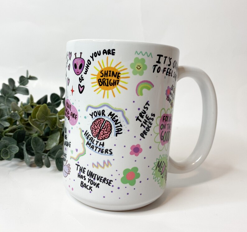 May include: White ceramic mug with a colorful design featuring positive affirmations and illustrations. The text includes "Shine Bright", "Your Mental Health Matters", "Trust the Process", and "The Universe Has Your Back".