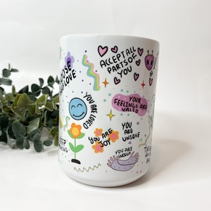 May include: White ceramic mug with colorful illustrations and positive affirmations, including "Accept all parts of you", "You are loved", "Your feelings are valid", "You are joy", "You are unique", and "You are enough".