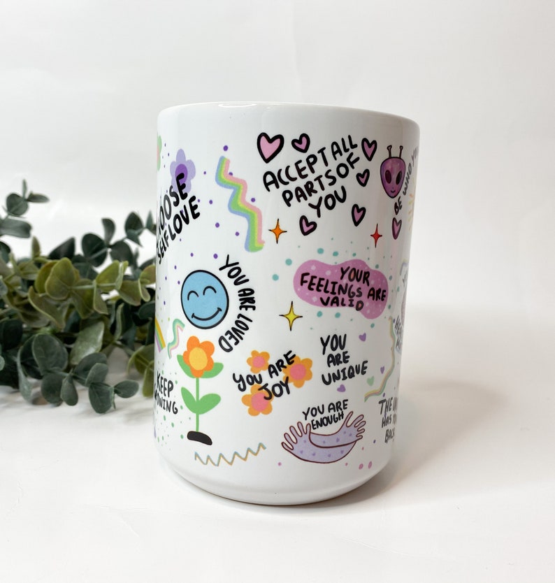 May include: White ceramic mug with colorful illustrations and positive affirmations, including "Accept all parts of you", "You are loved", "Your feelings are valid", "You are joy", "You are unique", and "You are enough".