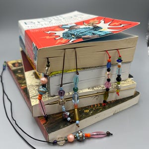 May include: A stack of books with several beaded bookmarks. The bookmarks feature various colorful beads, including pink, blue, and orange, strung on thin cords. The books have red and white covers.