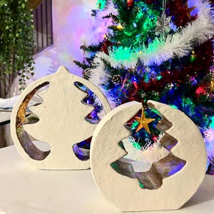 Peut inclure: Deux décorations de Noël en céramique blanche avec un motif découpé. Les décorations sont posées sur une surface blanche. La décoration de gauche a un accent de paillettes dorées. La décoration de droite a une étoile dorée sur le dessus.