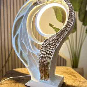 May include: A modern, abstract table lamp with a white base and a curved, textured design. The lamp features gold accents and a circular light source. The lamp is approximately 12 inches tall and has a power cord.
