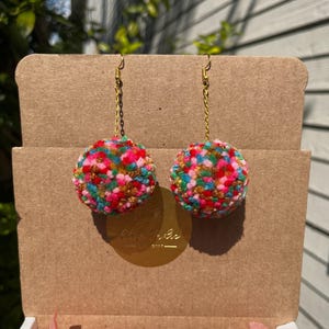 May include: A pair of colorful pom-pom earrings with gold chains and hooks. The pom-poms are made of yarn in shades of pink, green, blue, and yellow.