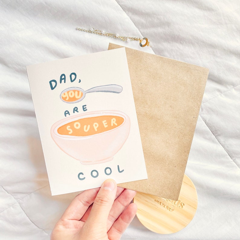 May include: A greeting card with the text "DAD, you are souper cool" and a bowl of soup illustration. The card is accompanied by a brown envelope, a wooden lid, and a delicate gold necklace with pearls.