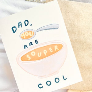May include: A greeting card with a whimsical illustration. The card features a spoon with the word "you" and a bowl of soup with the word "souper". The text "DAD, you ARE souper COOL" is written in blue and orange. The card is on a white surface.
