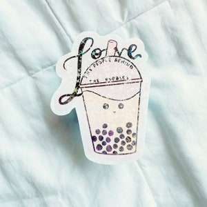 May include: A holographic sticker featuring a bubble tea design. The sticker has the word "Love" in cursive and the phrase "The people behind the bubbles" around the cup. The bubble tea has a smiling face and purple boba.