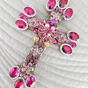 May include: Ornate cross pendant featuring a gold-tone base adorned with vibrant pink and fuchsia gemstones. The cross is embellished with floral details, pearls, and clear crystals, creating a luxurious and eye-catching design.