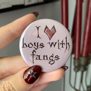 Pode incluir: Um botão branco com um coração rosa e texto preto que diz "I ♥ boys with fangs".