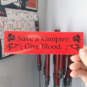 May include: Red sticker with black text and a black rose design. The text reads "Save a Vampire. Give Blood."
