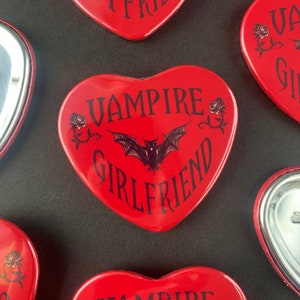 May include: Red heart-shaped pin with the text "Vampire Girlfriend" in black lettering. A black bat silhouette is in the center of the pin with two black roses on either side of the bat.