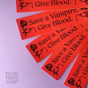 May include: Five red stickers with black text and a black rose graphic. The text on the stickers reads "Give Blood." "Save a Vampire. Give Blood." "Save a Vampire. Give Blood." "Save a Vampire. Give Blood." and "Save a Vampire. Give Blood."