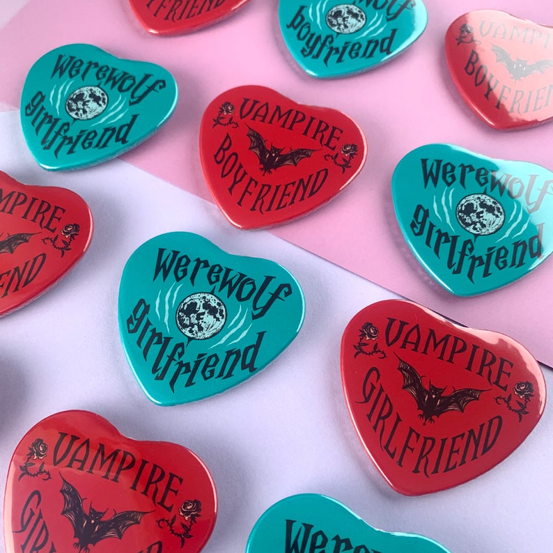 May include: A collection of heart-shaped pins with various designs featuring the words "Vampire Girlfriend", "Werewolf Girlfriend", and "Vampire Boyfriend". The pins are in red, blue, and turquoise.