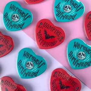 Peut inclure: Assortiment d'épingles rondes colorées sur le thème du loup-garou. Les épingles présentent une pleine lune, des chauves-souris et le texte "Werewolf Boyfriend" et "Werewolf Girlfriend".