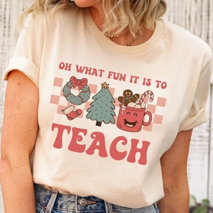 May include: A cream colored t-shirt with the text "Oh what fun it is to teach" in pink letters. The text is surrounded by a checkered pattern and Christmas themed illustrations.