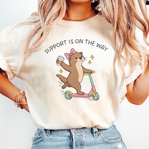 May include: An ivory t-shirt featuring a graphic of a brown otter wearing a pink bow, holding a coffee cup, and riding a pink scooter. The text above the otter reads SUPPORT IS ON THE WAY in black letters, accompanied by small yellow star accents.
