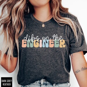 May include: A dark grey heather t-shirt with the text "dibs on the ENGINEER" in colorful, wavy letters.