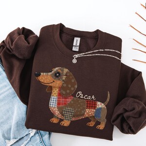 May include: A dark brown sweatshirt with a patchwork dachshund design. The dog's body is made of various fabric patterns, and the name "Oscar" is written below. A silver necklace with a pendant is draped across the sweatshirt.