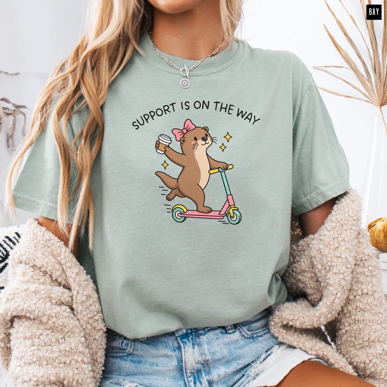 May include: A sage green t-shirt featuring a graphic of a brown otter wearing a pink bow, holding a coffee cup, and riding a pink scooter. The text above the otter reads SUPPORT IS ON THE WAY in black capital letters with small yellow star accents.