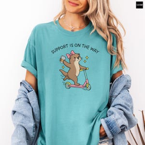 May include: A seafoam green t-shirt featuring a graphic of a brown otter wearing a pink bow, riding a pink and blue scooter while holding a coffee cup. The text above the otter reads SUPPORT IS ON THE WAY in black, arched capital letters.