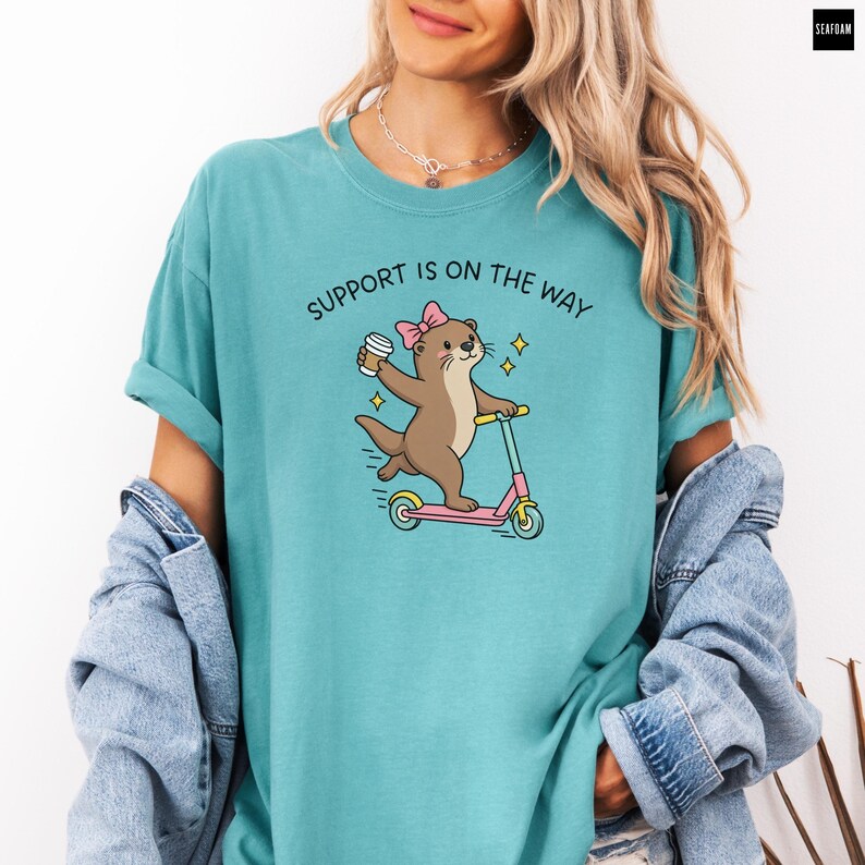 May include: A seafoam green t-shirt featuring a graphic of a brown otter wearing a pink bow, riding a pink and blue scooter while holding a coffee cup. The text above the otter reads SUPPORT IS ON THE WAY in black, arched capital letters.
