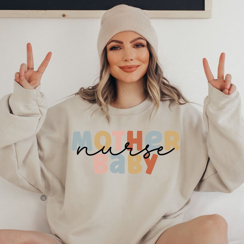 Pode incluir: Uma camisola bege claro com o texto "MOTHER nurse baby" em letras coloridas. As letras est&atilde;o em diferentes tons de rosa, azul, amarelo e laranja. O texto "nurse" est&aacute; escrito em it&aacute;lico preto.
