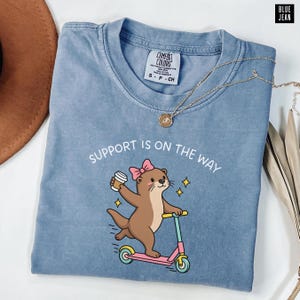 May include: A blue crew neck t-shirt featuring a graphic of a brown otter wearing a pink bow, holding a coffee cup, and riding a pink scooter. The text above the otter reads "SUPPORT IS ON THE WAY". The shirt has a Comfort Colors brand label.