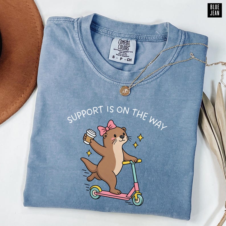 May include: A blue crew neck t-shirt featuring a graphic of a brown otter wearing a pink bow, holding a coffee cup, and riding a pink scooter. The text above the otter reads "SUPPORT IS ON THE WAY". The shirt has a Comfort Colors brand label.