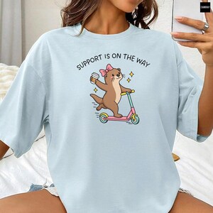 May include: A light blue t-shirt featuring a graphic of a brown otter wearing a pink bow, holding a coffee cup, and riding a pink and teal scooter. Above the otter, the text reads SUPPORT IS ON THE WAY in black capital letters.