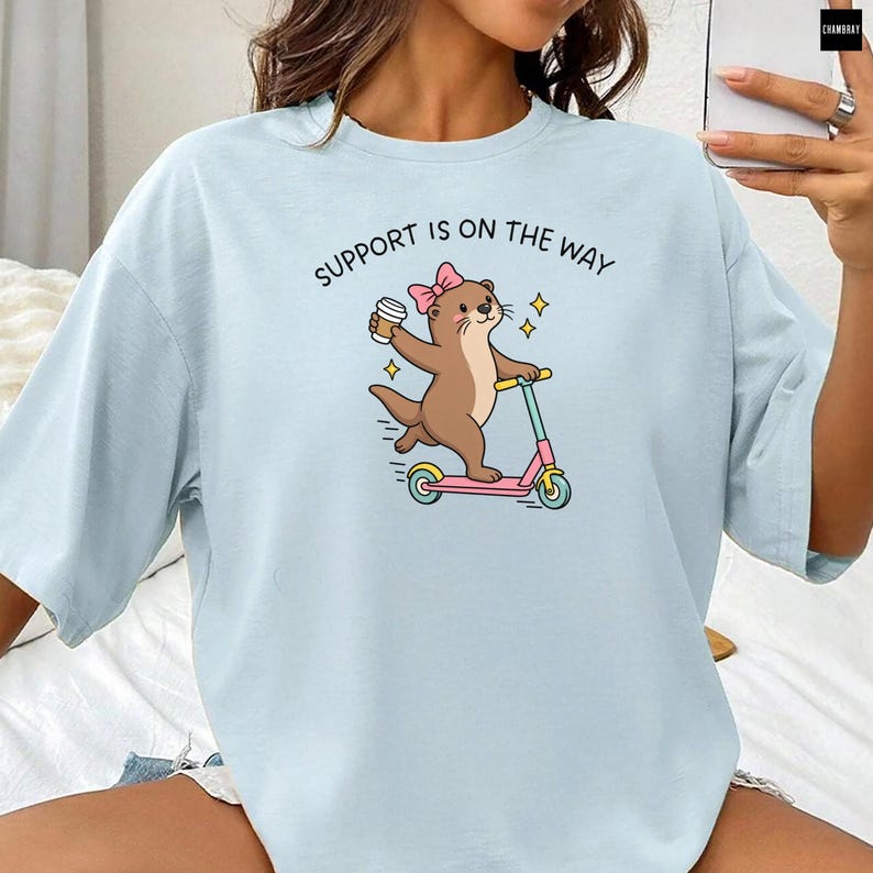 May include: A light blue t-shirt featuring a graphic of a brown otter wearing a pink bow, holding a coffee cup, and riding a pink and teal scooter. Above the otter, the text reads SUPPORT IS ON THE WAY in black capital letters.