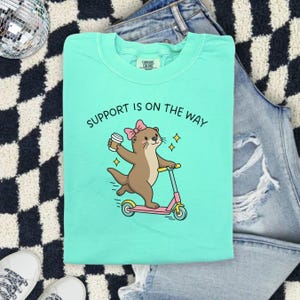 May include: A mint green crewneck t-shirt featuring a brown otter wearing a pink bow, holding a coffee cup, and riding a pink and yellow scooter. The text "SUPPORT IS ON THE WAY" is printed in black above the otter illustration.