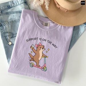 May include: A light purple t-shirt featuring a brown otter wearing a pink bow, riding a pink and blue scooter while holding a coffee cup. The text above the otter reads SUPPORT IS ON THE WAY in black letters. The shirt has a Comfort Colors label.