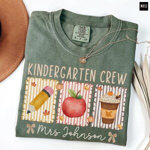 May include: A moss green t-shirt with the words "KINDERGARTEN CREW" and "Mrs Johnson" printed on the front. The design includes a pencil, apple, and coffee cup with fall-themed accents. A silver necklace is draped across the shirt.