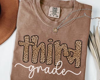 May include: A brown Comfort Colors t-shirt with the words "third grade" in a combination of leopard print and white script. A silver necklace with a small pendant rests on the shirt. A brown hat is visible in the upper left corner.