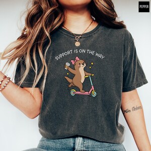 May include: A charcoal gray short-sleeve t-shirt featuring a graphic of a brown otter wearing a pink bow, holding a coffee cup, and riding a pink and blue scooter. The text above the otter reads, "SUPPORT IS ON THE WAY" in white letters.
