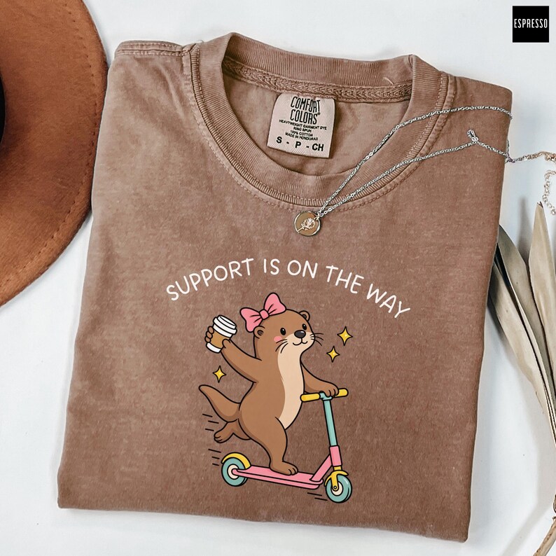 May include: A brown t-shirt featuring a graphic of an otter with a pink bow riding a pink scooter while holding a coffee cup. The text above the otter reads SUPPORT IS ON THE WAY. The shirt has a crew neckline and a Comfort Colors brand label.