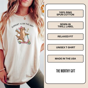 May include: A cream-colored t-shirt featuring a graphic of a brown otter with a pink bow riding a pink scooter while holding a coffee cup. The text reads SUPPORT IS ON THE WAY. The shirt details include 100% ring spun cotton, a sewn-in twill label, relaxed fit, unisex design, and made in the USA.