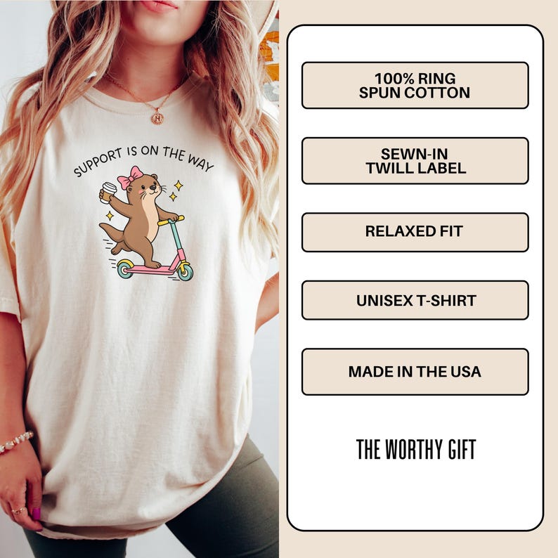 May include: A cream-colored t-shirt featuring a graphic of a brown otter with a pink bow riding a pink scooter while holding a coffee cup. The text reads SUPPORT IS ON THE WAY. The shirt details include 100% ring spun cotton, a sewn-in twill label, relaxed fit, unisex design, and made in the USA.